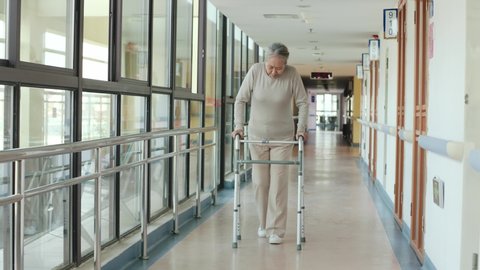 senior asian woman walking slowly with help of a zimmer frame in corridor of nursing home