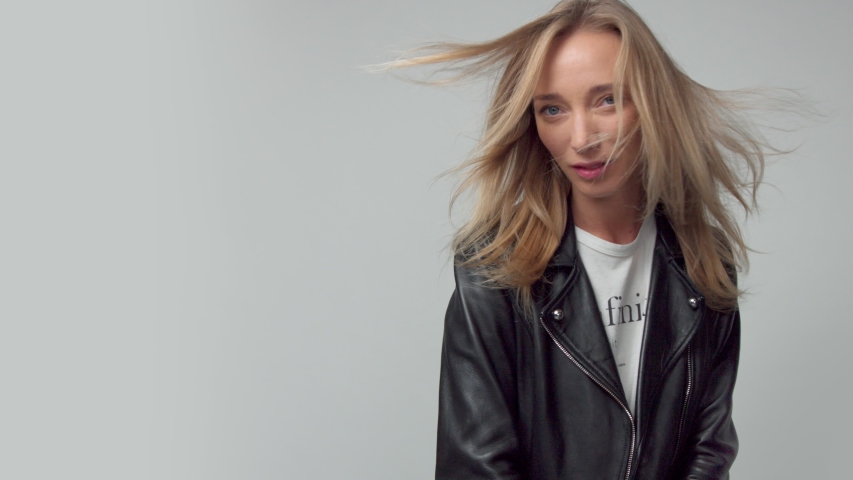 panned portrait of blonde woman with hair blowing, in studio alone, wears a leather jacket