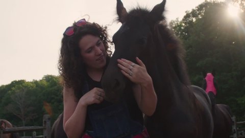 Young woman farmhand greets & pets a family of horses in meadow during evening