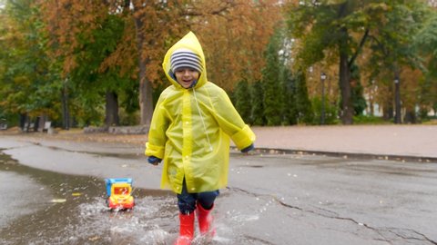 4k slow motion footage of happy smiling little boy pulling toy truck and running over puddles at autumn park after rain