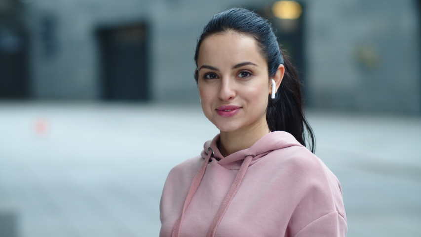 Close up happy woman listening music in earphones outdoor. Portrait of cheerful woman posing for camera in wireless earphones. Joyful girl smiling into camera in slow motion.