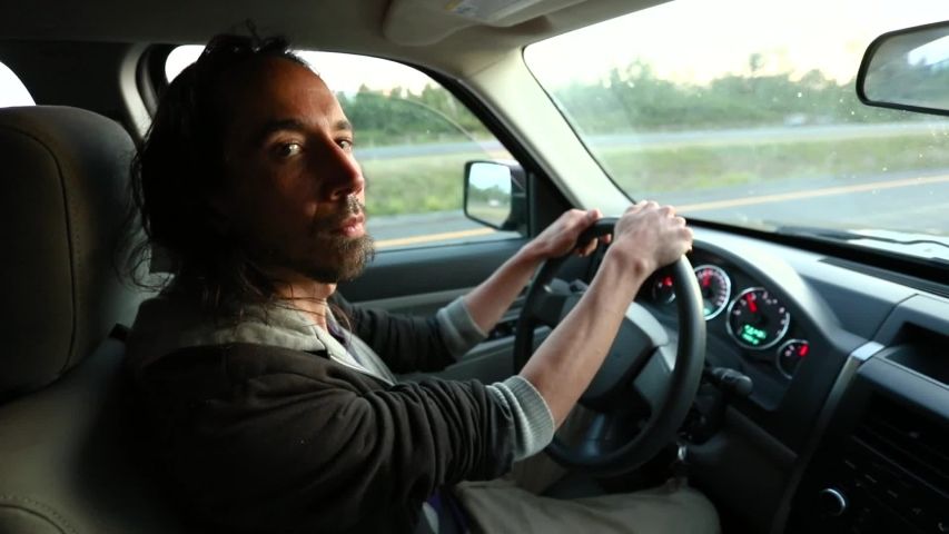 A man with long hair and beard is driving on a highway in the afternoon. He looks to camera, raises his eyebrows and smiles with hands on the wheel 
