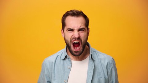 Angry swearing unshaven bearded young guy 20s in denim shirt white t-shirt isolated over yellow background in studio. People sincere emotions, lifestyle concept. Looking at the camera scream and shout