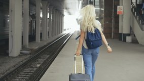 Back view of young positive tourist girl walking and dragging luggage suitcase searching for her train on platform. Caucasian blonde woman waiting for train and planning happy holiday vacation.