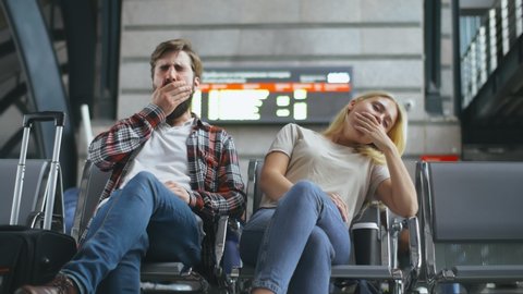 Tired bored young caucasian couple sitting and yawning in departure lounge with suitcase and waiting for delayed flight. Stressed and exhausted tourists waiting for their train at railway station