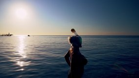 Woman wearing a snorkel entering a calm sea backlit by a hot sun casting a reflection over the water in a summer vacation concept