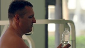 Shirtless man taking a drink of water from a plastic bottle while working out in a gym in a close up side view