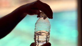 Person opening a plastic bottle of bottled water for a drink alongside a sparkling blue pool in a close up on the hand