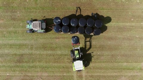 Aerial top down view of tractors working together picking up plastic wrapped bales with front pickup arm the silage is fermented high-moisture stored fodder which can be fed to cattle during winter 4k