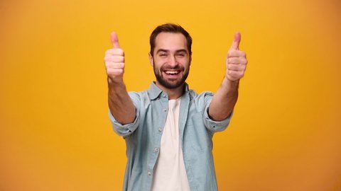 Handsome smiling bearded young guy 20s in denim shirt white t-shirt isolated on yellow background in studio. People sincere emotions, lifestyle concept. Looking approvingly at camera showing thumbs up