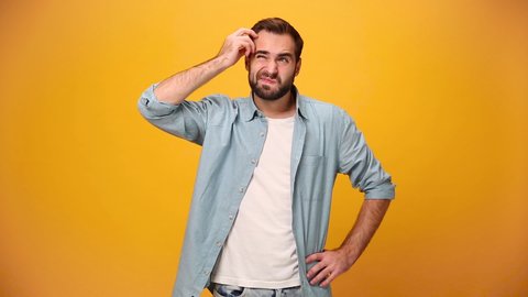 Bearded young guy 20s in denim shirt white t-shirt isolated on yellow background in studio. People sincere emotions, lifestyle concept. Looks at camera, thinks, scratches at temple comes up with ideas