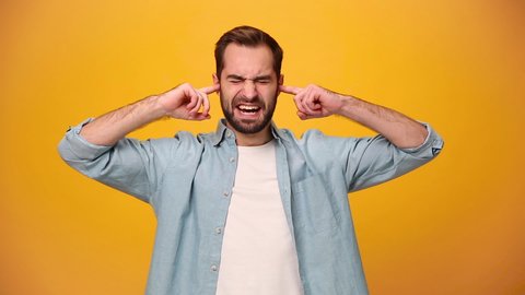 Angry bearded young guy 20s in denim shirt white t-shirt isolated over yellow background in studio. People sincere emotions, lifestyle concept. Looking at the camera. Covering ears with fingers scream