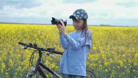 Girl with camera in yellow field. A cute child in a cap makes photos of canola field by a bike.