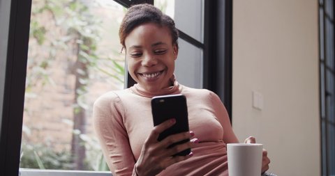 Close up low angle view of happy young African woman sitting in sofa looking at mobile phone black young girl enjoy coffee at weekend morning in cozy home with good mood