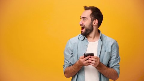 Handsome unshaven bearded young guy 20s in denim shirt white t-shirt isolated over yellow background in studio. People sincere emotions, lifestyle concept. Looking camera wow aside using mobile phone