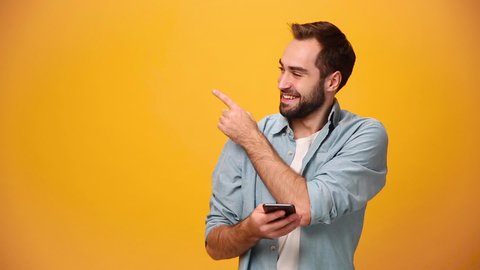 Handsome unshaven bearded young guy 20s in denim shirt white t-shirt isolated on yellow background in studio. People sincere emotions lifestyle concept. Pointing aside on copy space using mobile phone