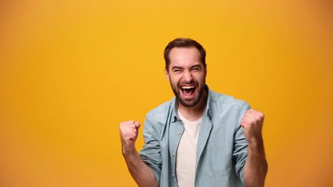 Handsome bearded young guy 20s in denim shirt white t-shirt isolated over yellow background in studio. People sincere emotions, lifestyle concept. Looking at the camera doing winner gesture, say yeah