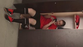 Vertical format video of young female athlete in soccer uniform sitting in locker, typing on smartphone and chatting with friend before training