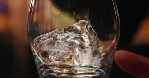Macro of a professional bartender is putting professional crystal ice cubes in a glass while preparing an alcoholic cocktail to customers at the bar or disco club.