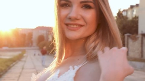 Pretty looking Woman Standing in front of Camera at Sunset, touching her Blonde hair. Looking gorgeous and Cute. Posing like a model. Smiling at Camera. Having Beautiful eyes.