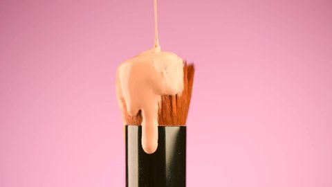 Makeup liquid foundation pouring on make-up contouring brush, closeup. Foundation beauty facial cosmetics, tool for perfect make up. Dripping bb cream or concealer, over pink background. 4K slow mo