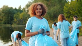 Smiling Afro-American volunteer holding bin bag with trash on polluted lake shore while multi-ethnic team of eco activists is collecting garbage working together. - Powered by Shutterstock - Get 15% off with code: PIKWIZARD15