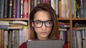 A young woman is reading a book in a tablet. A woman with glasses carefully looks at the tablet. In the background are books on bookshelves. Book library. - Powered by Shutterstock - Get 15% off with code: PIKWIZARD15