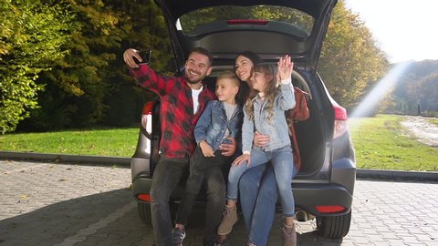 Front view of cheerful lovely parents with cute teen kids which making selfie with waving hands,sitting in the car's trunk
