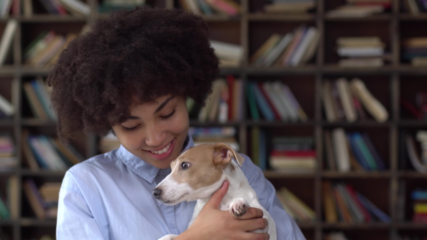 Young african woman standing in library hugging dog stroking happy smiling happy close-up