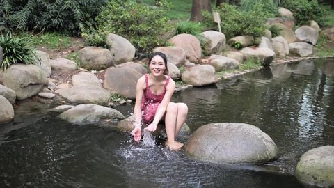 Sexy Chinese young woman in red dress sitting on big rocks in river and playing with water, barefoot girl enjoy water in summer, super slow motion footage, summer concept.