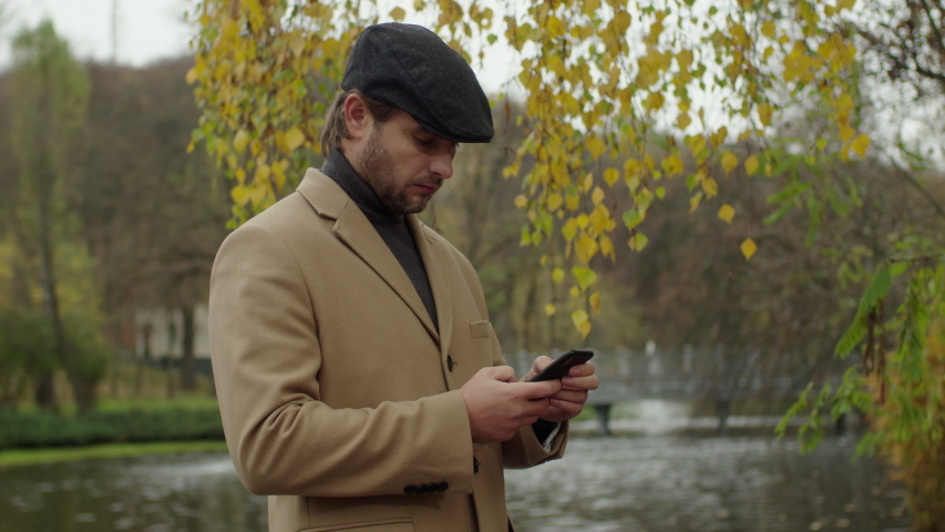 Bearded man wearing classical clothes typing a message on his smartphone against the backdrop of an incredibly beautiful autumn landscape