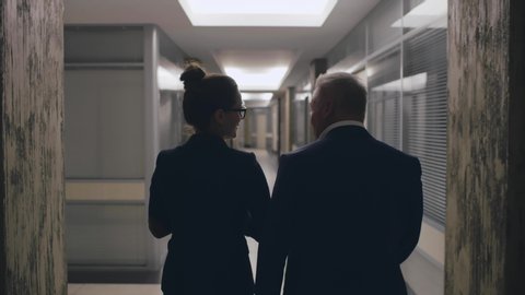 Young businesswoman going along long narrow corridor with businessman in formal suit back view pov camera sliding. Partners in business suit clothing walk to meeting work indoors. Office style success