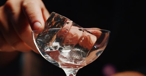 Close up of a bartender is putting professional crystal ice cubes in a glass while preparing an alcoholic cocktail to customers at the bar or disco club.