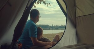 Happy bonding traveler father and son sitting near tent camp around mountains lake under sun light enjoying the leisure and freedom. 4K slow motion video