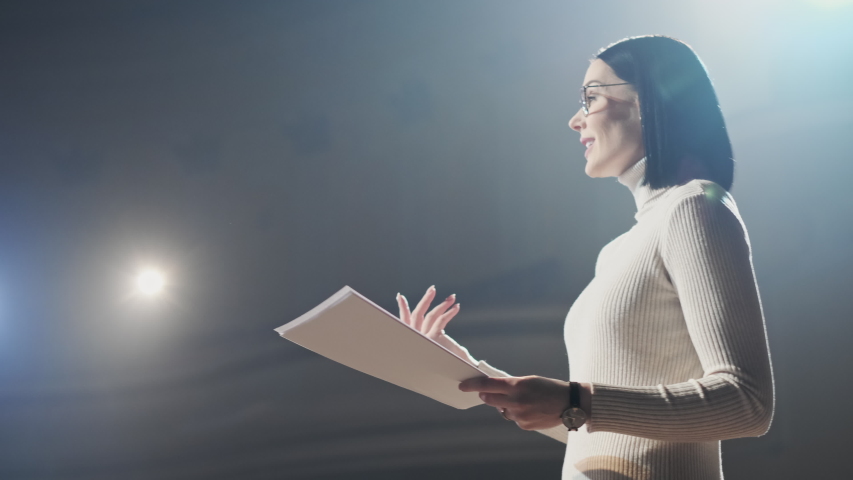 Adult female speaker with short hair in a grey dress standing on stage and reporting for audience. Expert perform on the stage address to modern crowd audience sitting and listening in large hall. Royalty-Free Stock Footage #1043066002