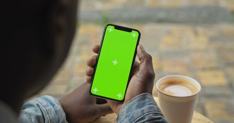 Lviv, Ukraine - August 09, 2019: Close Up Shot of Afro American Man Sitting at Table with Paper Coffee Cup Holding his Smartphone with Green Screen and Looking on it Scrolling Touching Screen Backside
