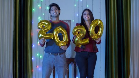 Pan shot of happy Indian couple celebrating Christmas or New Year together at home. Adorable young friends holding and dancing with golden 2020 balloons during New Year and Christmas time in India