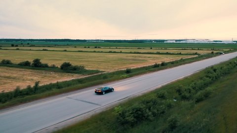 Black car driving down an asphalt road crossing the field on summer day. Russia Saint-Petersburg 20.07.2019