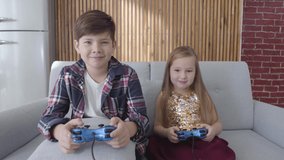 Two Caucasian children playing video game as sitting on couch at home. Boy and girl winning, making victory gesture and hugging. Leisure, childhood, happiness.