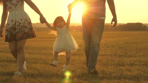 happy little daughter holds mom and dad hands and jumps on meadow in park in rays of yellow sun. Family concept. child plays with dad and mom on field in sunset light. Walking with small kid in nature