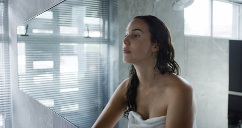 Side view close up of a young Caucasian woman with long dark hair wearing a bath towel looking in the mirror and massaging her face with her fingers in a modern bathroom, slow motion