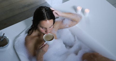 High angle view of a young Caucasian woman with long dark hair relaxing in a foam bath with lit candles beside the bath, drinking a cup of coffee, slow motion