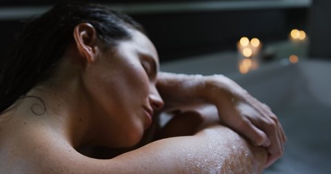 Close up side view of a young Caucasian woman with long dark hair relaxing in a foam bath, leaning and resting her head on her arms with eyes closed and smiling, lit candles in the background, slow