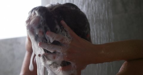 Rear view head shot of a young Caucasian woman with long dark hair standing under the shower in a modern bathroom, washing and rinsing her hair, slow motion