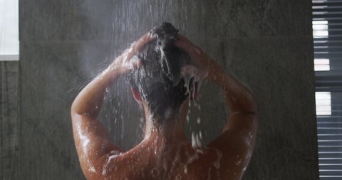 Rear view close up of a young Caucasian woman with long dark hair standing under the shower in a modern bathroom, washing and rinsing her hair, slow motion