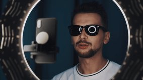 Active man video logger with dark hair, in white t-shirt and wearing sunglasses, is speaking, looking at camera of smartphone in front of ring light, shooting video for blog. Shot on 4K RED camera.