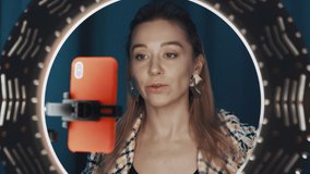 Young cute female fashion blogger sits in front of ring light and orange smartphone, adding blush to her face, showing technique of makeup, streaming tutorial beauty video. Shot on 4K RED camera.