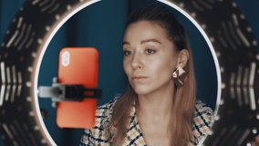 Pretty female beauty blogger sits in front of ring light and orange smartphone, doing contouring on her face, showing technique of makeup, recording tutorial beauty video. Shot on 4K RED camera.