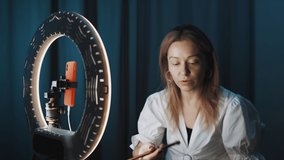 Young female fashion blogger with blond hair holds mirror and puts shadows on eyes with brush, sitting in front of ring light, streaming online whole process. Shot on 4K RED camera