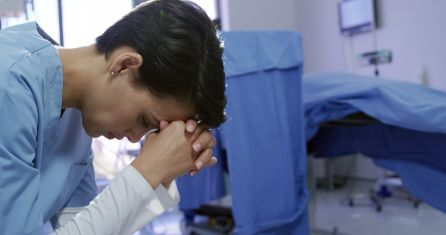 Mixed race female healthcare professional wearing scrubs in a hospital operating theatre, sitting with head in hands in despair. Healthcare workers in the Coronavirus Covid19 pandemic | Shutterstock HD Video #1043121754